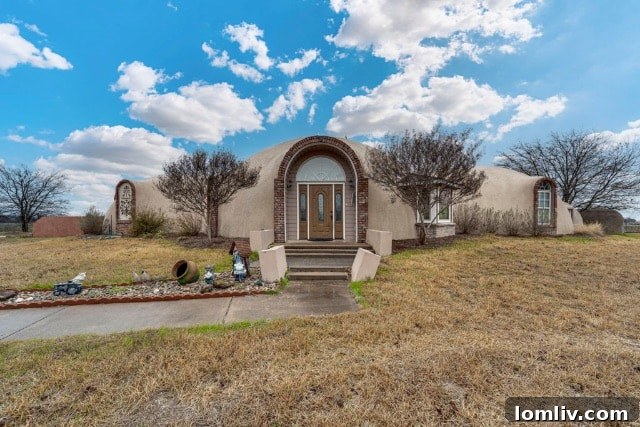 An aerial view of the Monolithic Dome Institute complex, featuring various dome structures and a large caterpillar-like building, set against a backdrop of green fields.