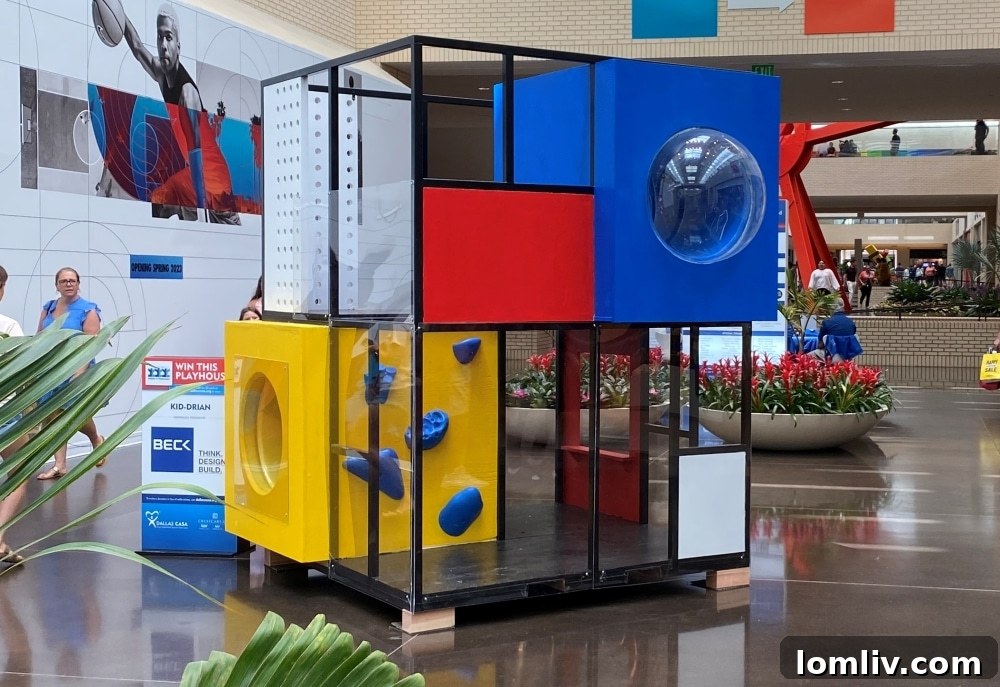 A vibrant, avant-garde playhouse design featuring geometric shapes and bold colors, set against a backdrop of a public space, possibly NorthPark Center, with people in the background.
