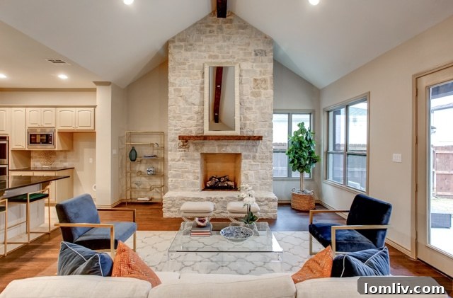 An open-plan living space showing a large kitchen transitioning into a family room with a beamed ceiling and a prominent stone fireplace.